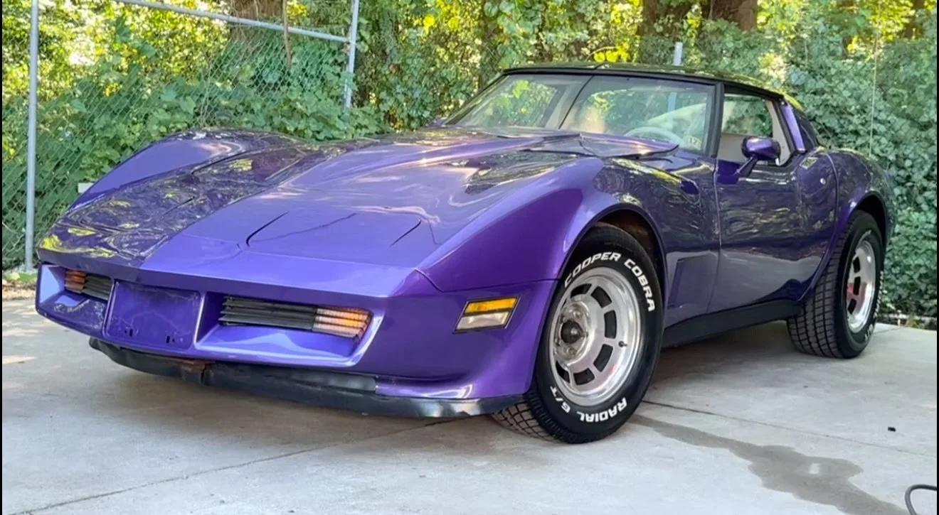 A purple sports car parked on a concrete surface with trees and a metal fence in the background.