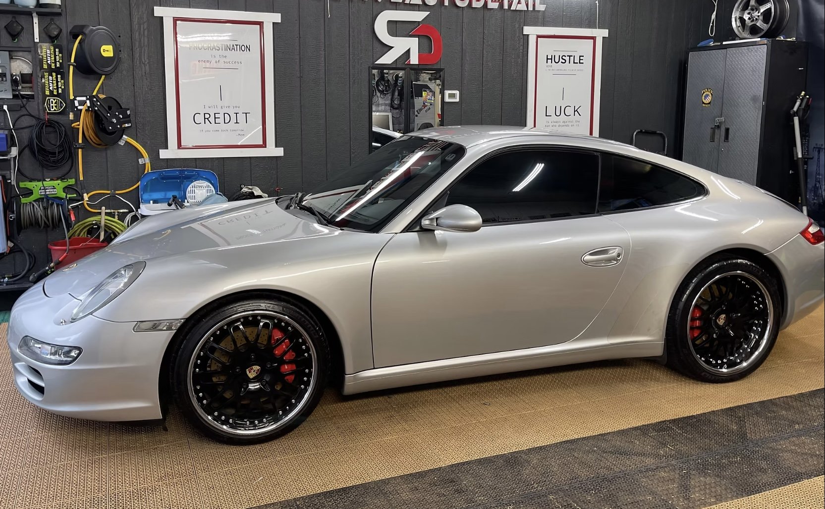 A silver Porsche sports car parked in a garage with various tools and signs on the wall, including motivational posters and project logos.