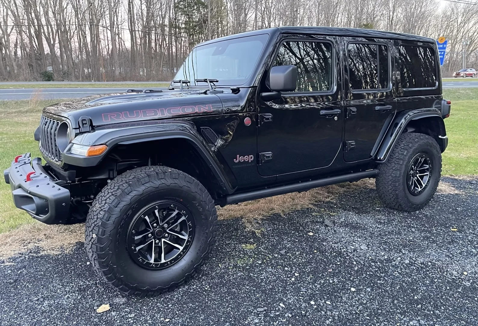 Black Jeep Wrangler Rubicon parked on a gravel surface, with leafless trees in the background.