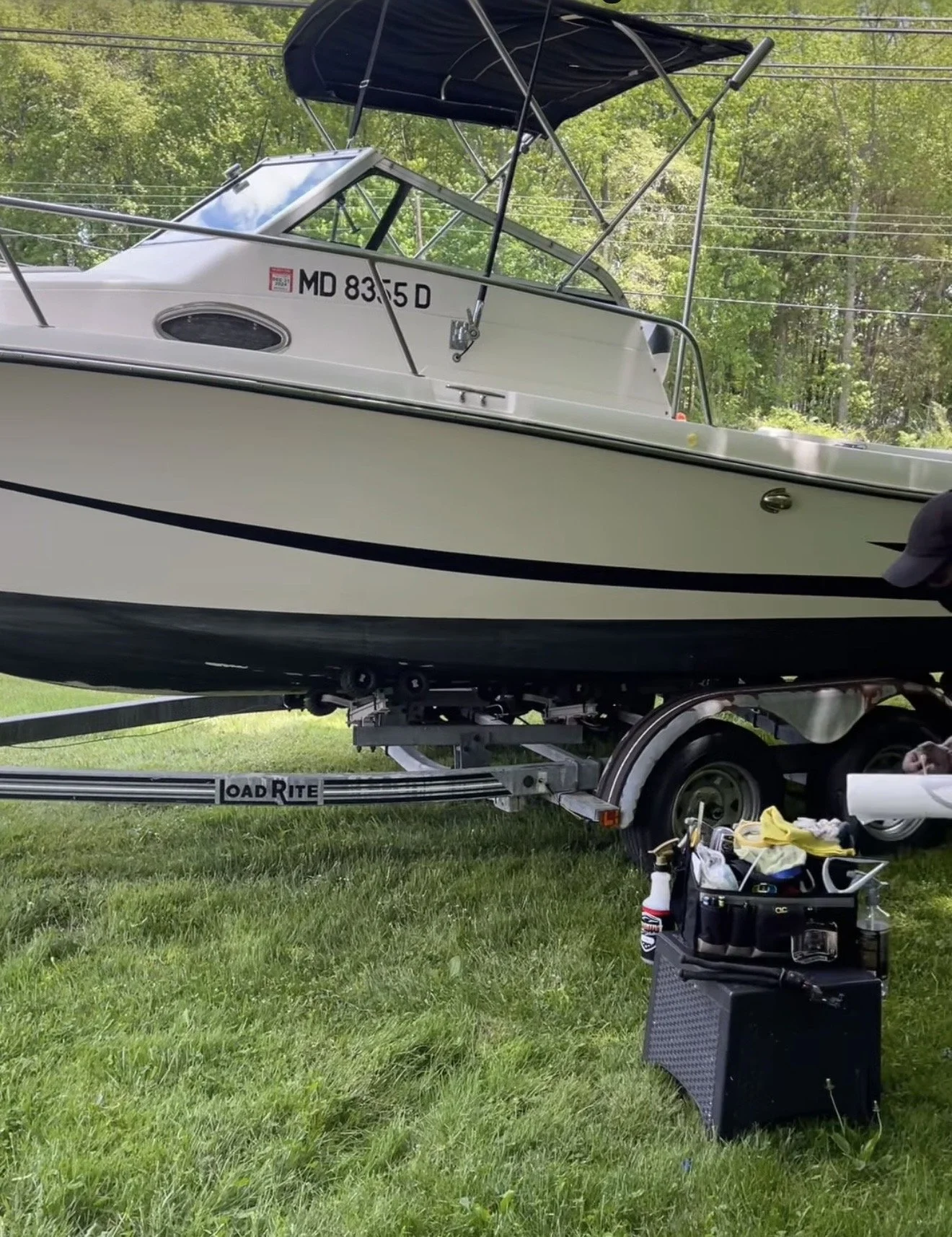 A white boat on a trailer, parked on a grassy area with trees in the background. The boat has a canopy and the trailer has a toolbox and some tools and supplies nearby.
