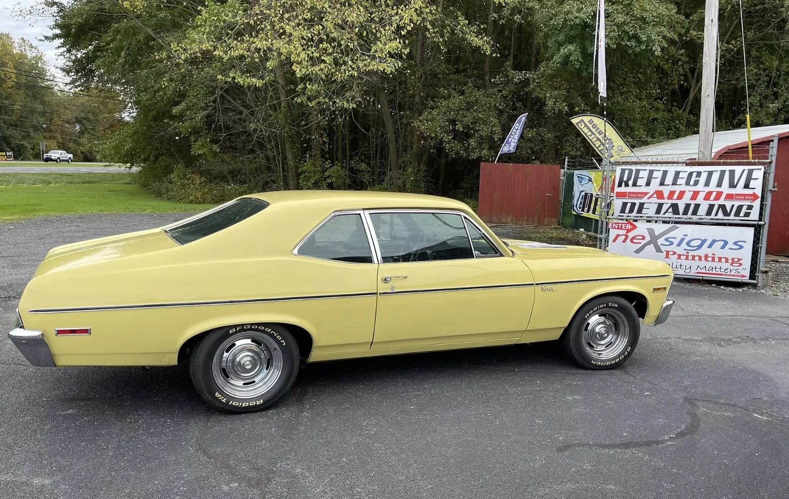 A yellow vintage car parked on a paved lot with a backdrop of trees, next to a sign reading Reflective Auto Detailing, with flags and a fence.