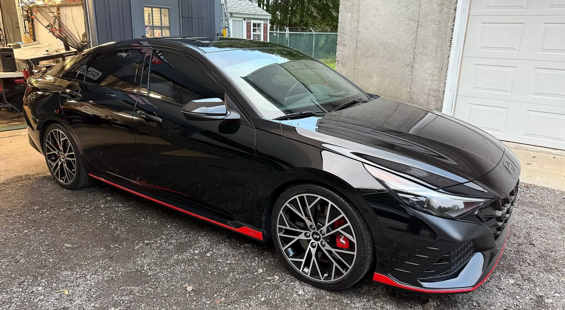 Black sports car with red accents parked in a residential driveway beside a garage with a closed white door. The car features angular headlights, tinted windows, and multi-spoke wheels with a red brake caliper visible behind the front wheel.