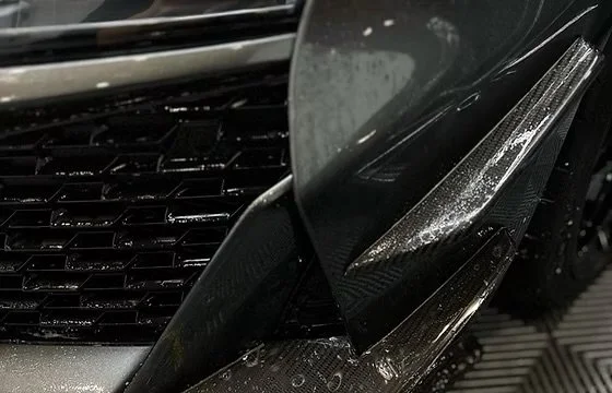 Close-up of a black car's front end featuring a carbon fiber front splitter and a glossy black grille.