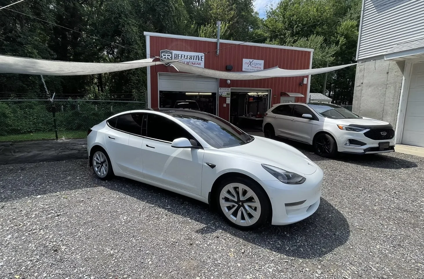 Two white cars parked outside a red and gray metal building with open garage doors, surrounded by trees and a gravel lot.