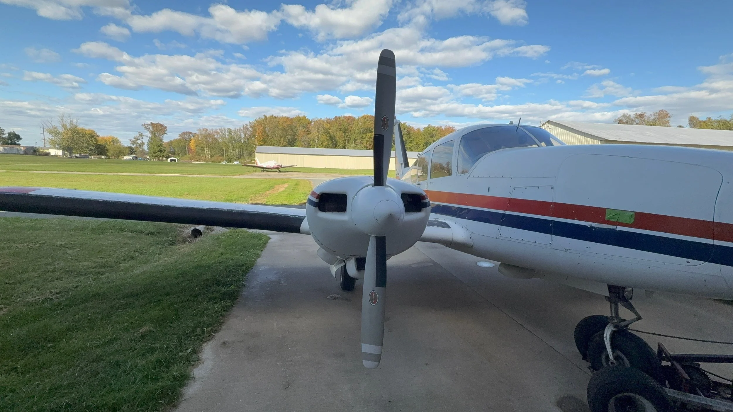 Small propeller airplane parked on the tarmac with a grassy field and hangars in the background, under a partly cloudy sky.