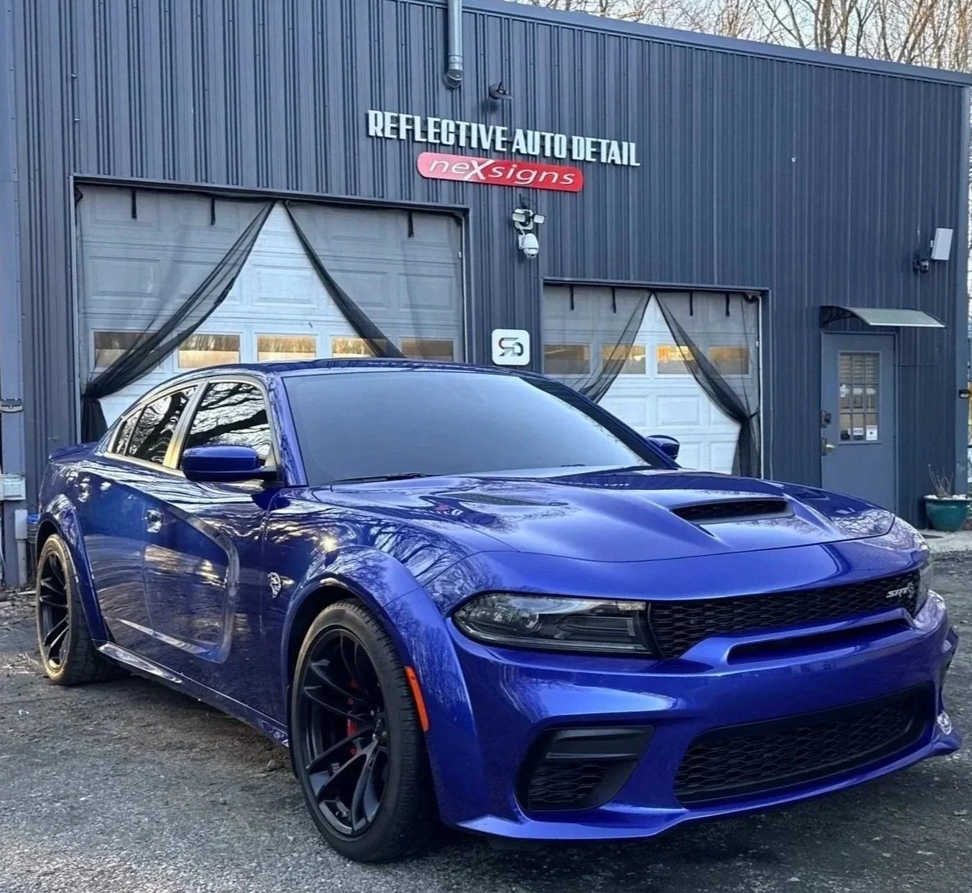 A blue sports car parked outside a dark blue auto detailing shop with sign reading 'Reflective Auto Detail' and 'nex signs.' The shop has two garage doors with black curtains and a small side door to the right. Trees are visible in the background.