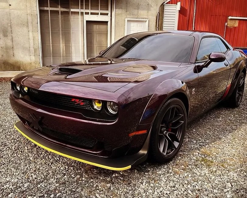 A purple Dodge Challenger SRT with yellow accents, parked on gravel in front of a building with concrete and red siding.