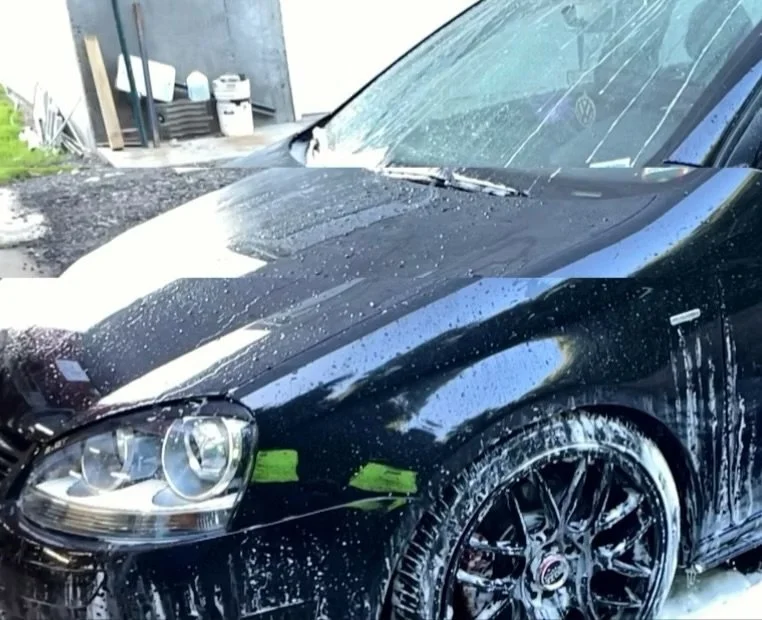 Black Volkswagen with water droplets and soap suds on the hood and front fender, with a wet windshield, parked in a driveway or car wash area.