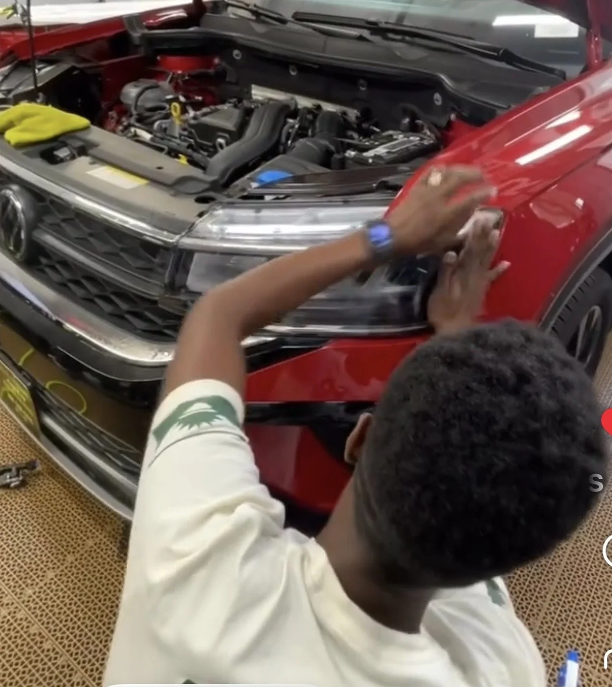 A person working on a red car with the hood open, inspecting the engine. The person is wearing a white shirt with a green logo on the sleeve and a blue watch.
