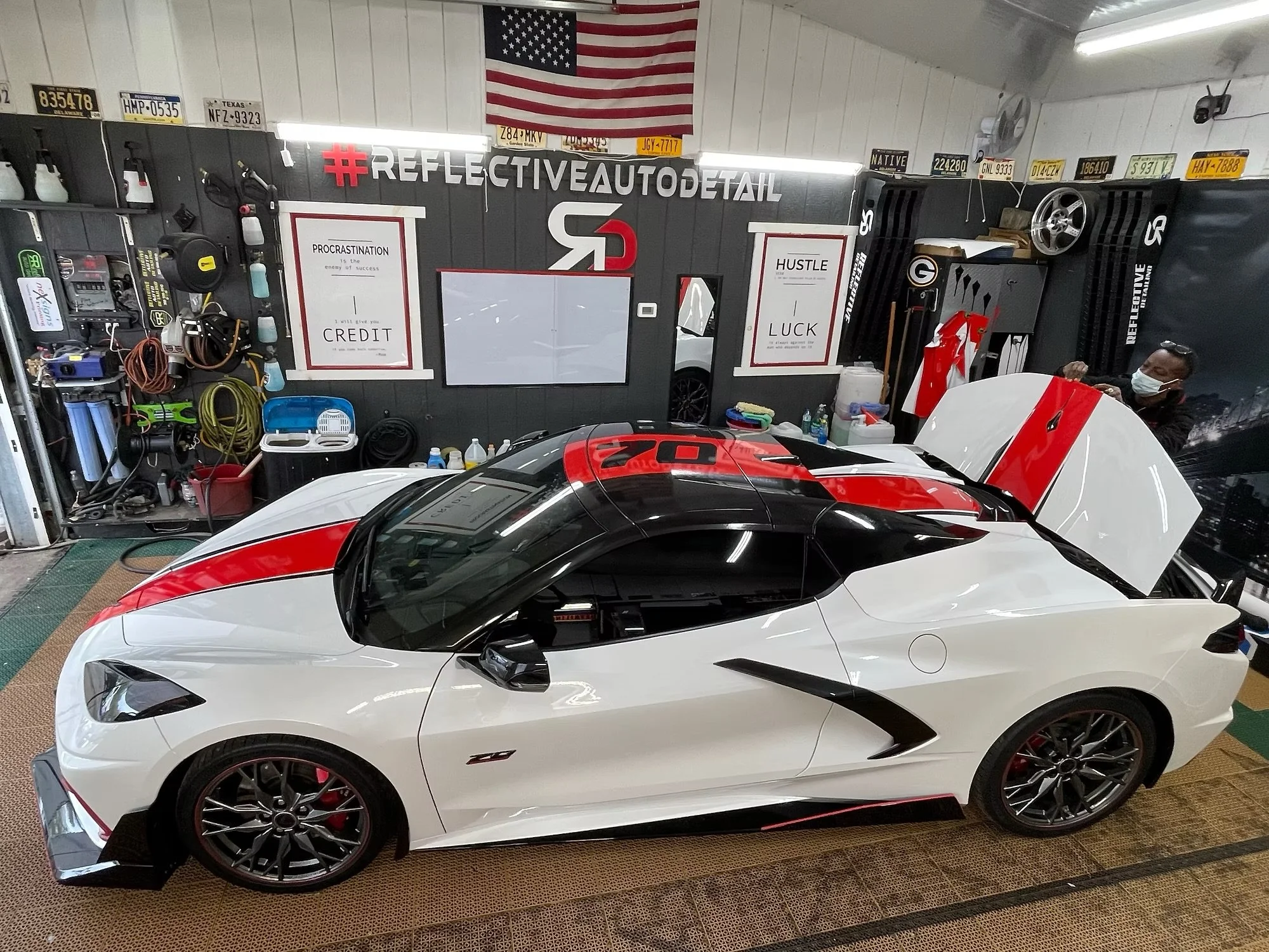 A white sports car with red and black accents parked inside a garage. The garage wall has various tools, equipment, and decorations, including a large hashtag #REFLECTIVEAUTODETAIL and American flags. There is a person wearing a face mask working on 