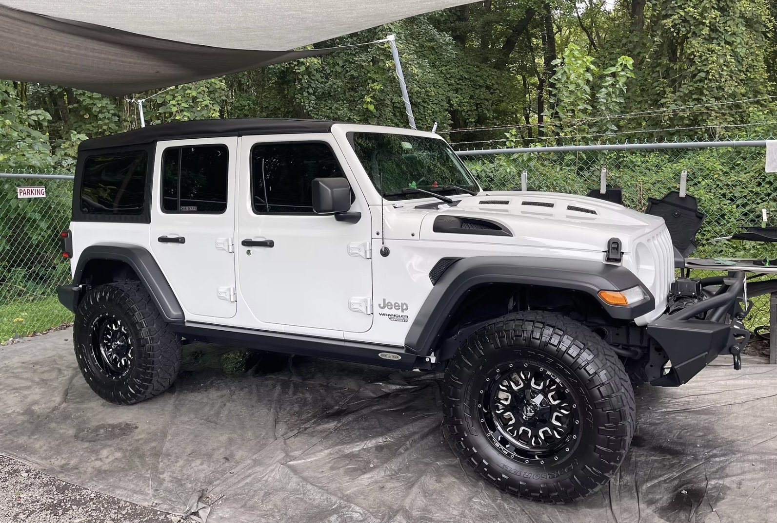A white Jeep Wrangler with off-road tires parked on a concrete surface in front of a chain-link fence with trees behind. The vehicle has a black front bumper and fender flares.
