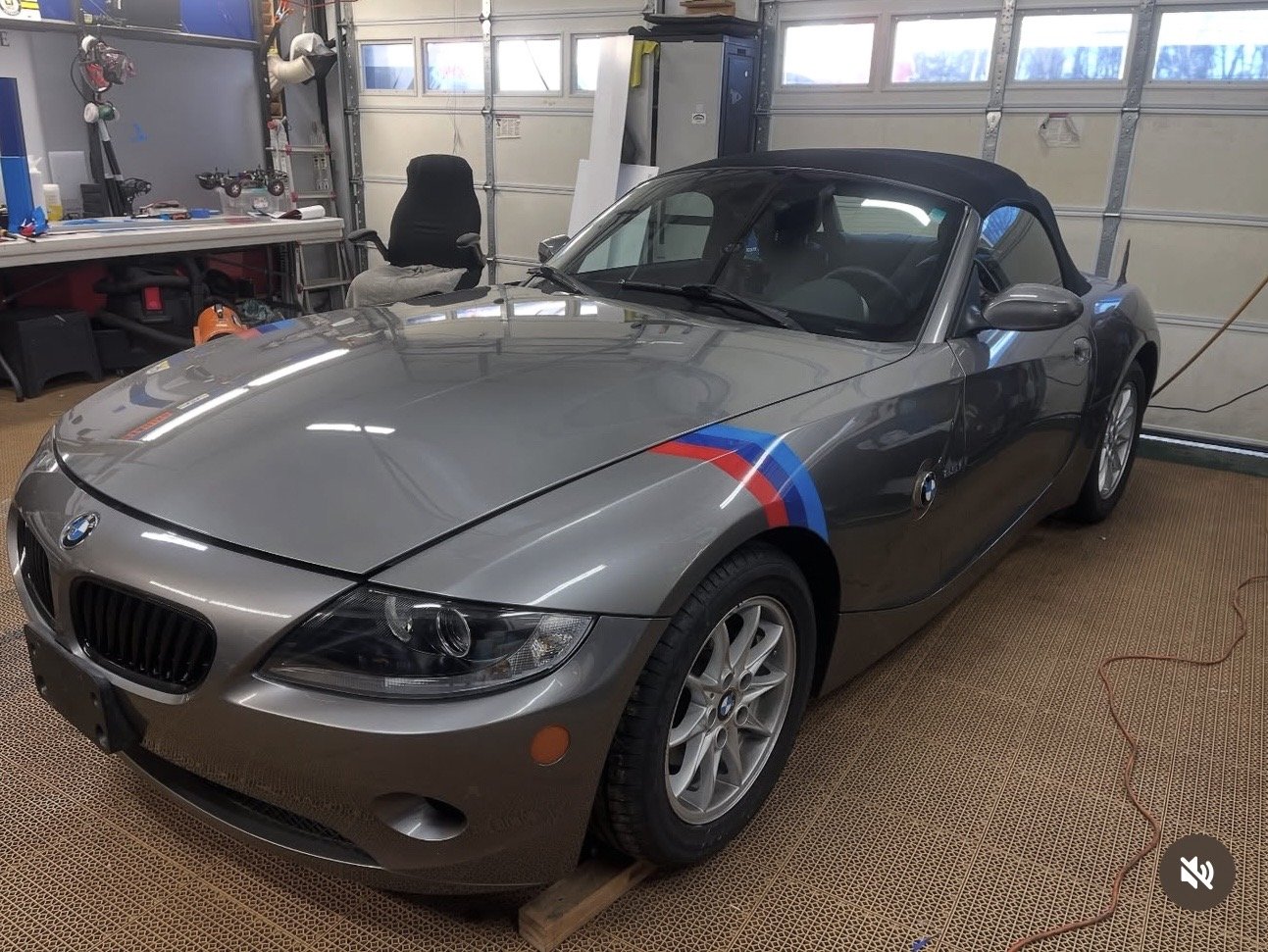 A silver BMW Z4 convertible with a black soft top, parked inside a garage. The car has BMW Motorsport stripes in red and blue on the front fender.