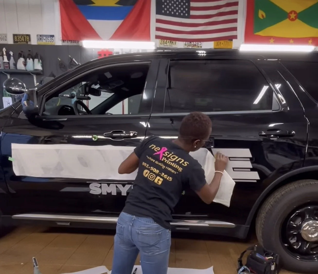 A woman is installing custom graphics on a black SUV inside a workshop. Flags from various countries are hanging overhead. The woman is wearing a black t-shirt with business contact information and is focused on applying decals to the vehicle's side.