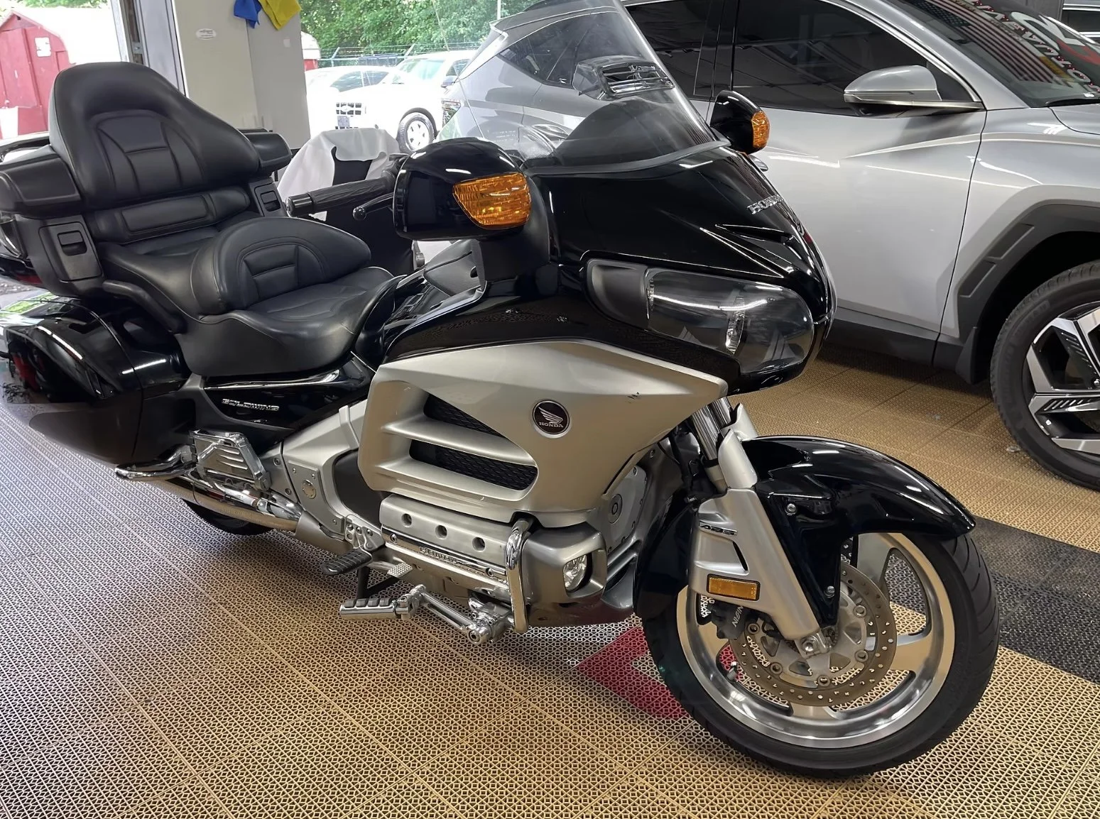 A gold and black Honda Gold Wing touring motorcycle parked indoors next to two modern cars, with a parking lot visible through the window behind.
