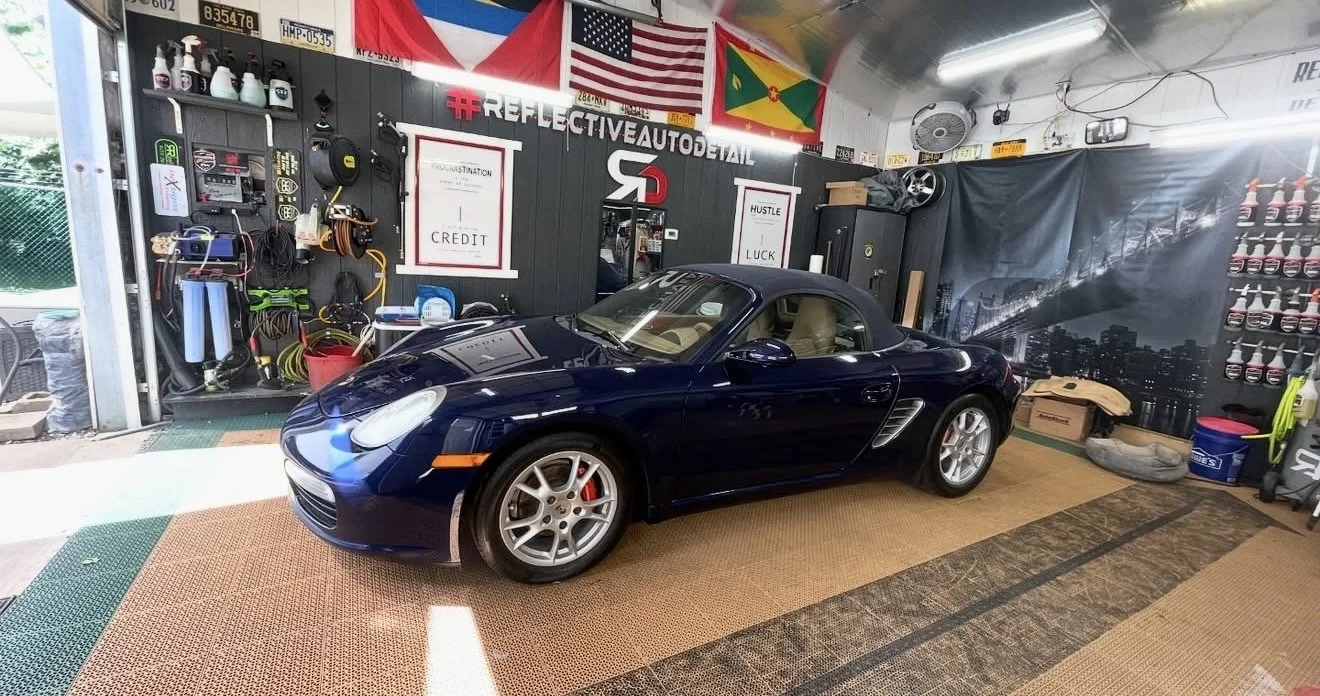 A dark blue Porsche convertible parked inside a garage with various tools, equipment, and car care products on the walls. Flags of the United States, Grenada, and the United States Navy hang above. The garage has a cityscape backdrop on one wall and 