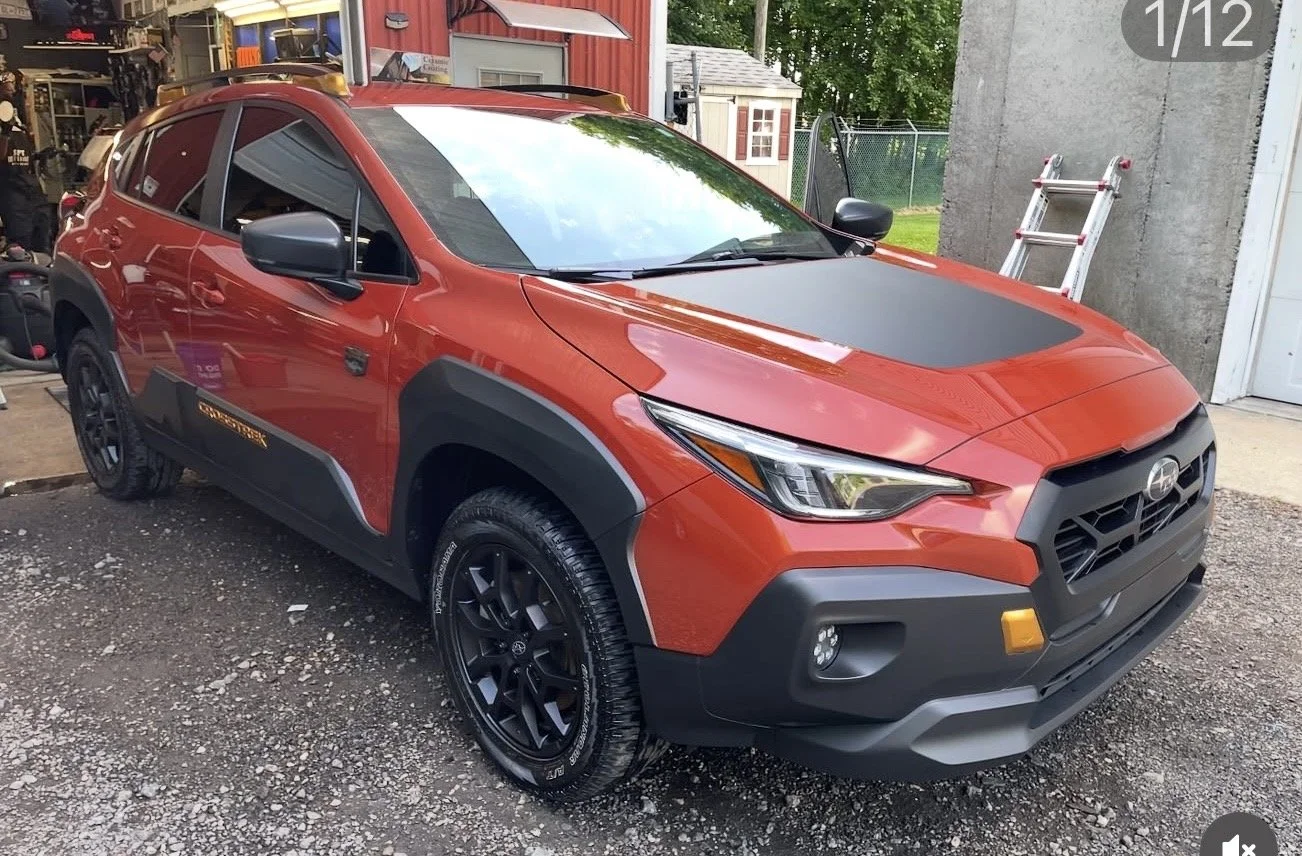 A red Subaru Crosstrek parked outside near a garage, with a black hood and black wheels, reflecting the surroundings in its windows and windshield.