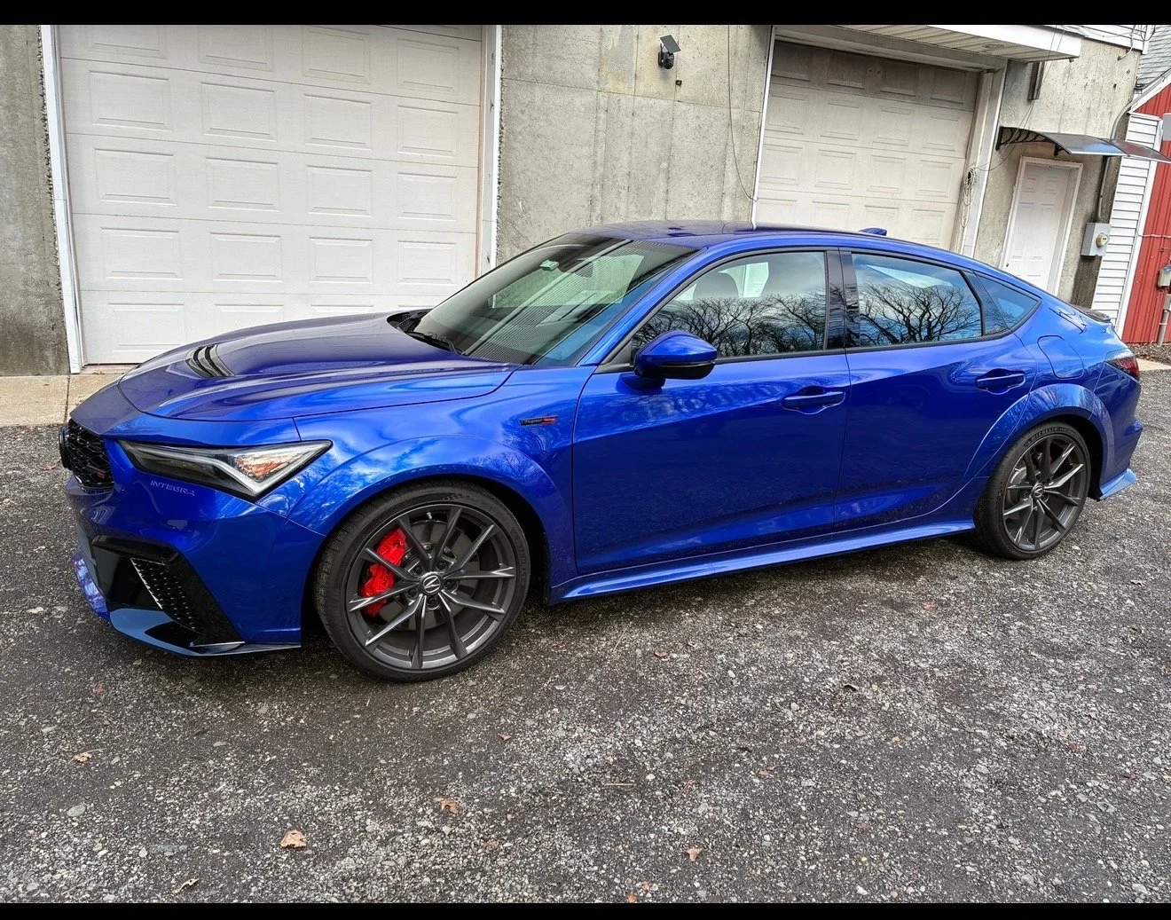 A blue sports car parked in front of two closed garage doors on a gravel driveway.
