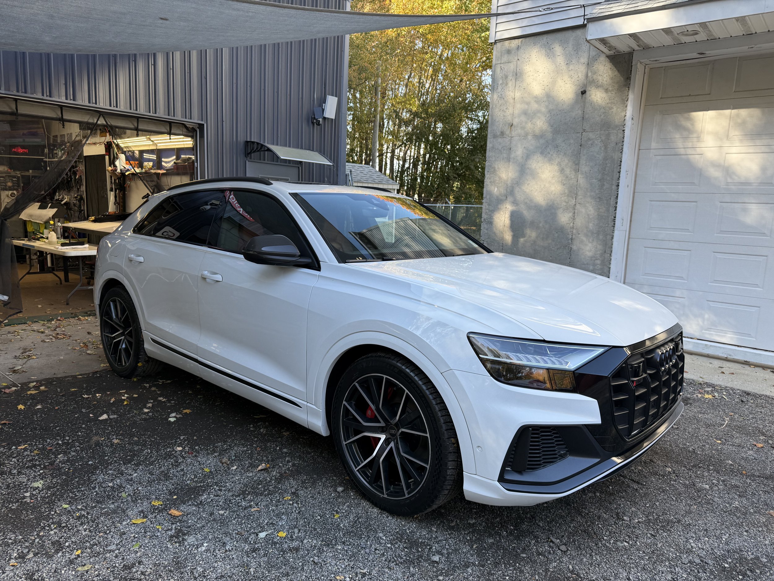 A white Audi sports car parked in a driveway, with a garage and trees in the background.
