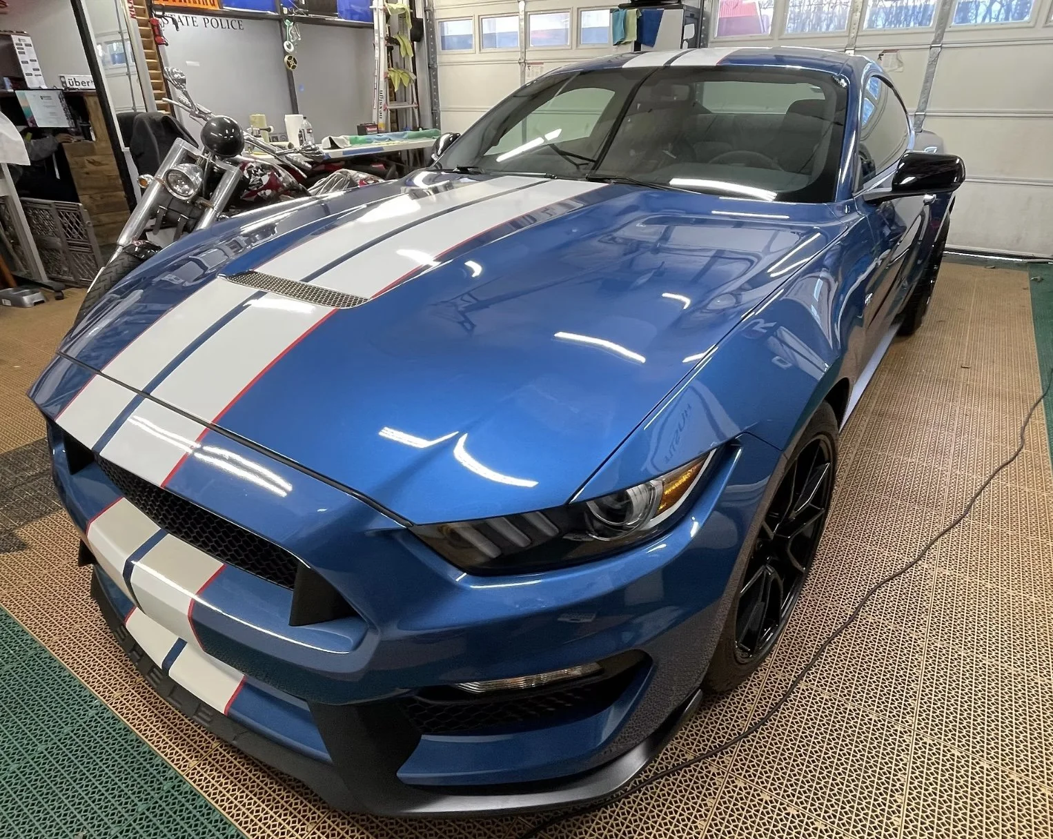 A blue Mustang with white racing stripes parked in a garage. The car has sleek, modern design with black wheels and is positioned on a rubber mat. There are various items and tools on shelves in the background.