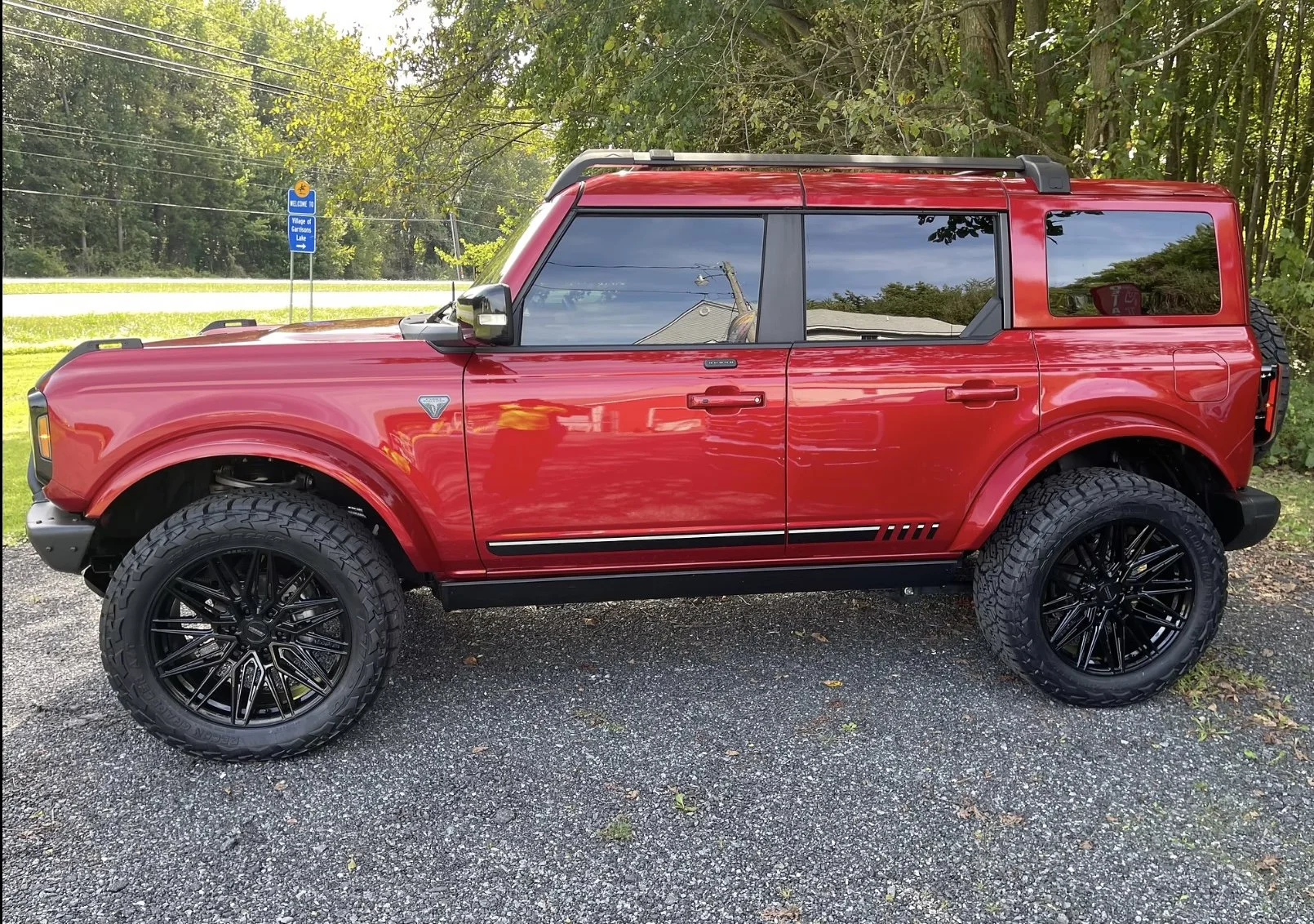 Red off-road vehicle with large tires and black rims parked on gravel ground, surrounded by greenery with trees and a blue sign in the background.