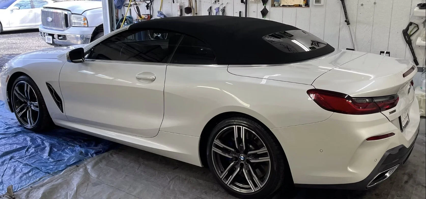 A white BMW convertible with a black soft top inside a garage, parked on a blue tarp with another vehicle visible outside.