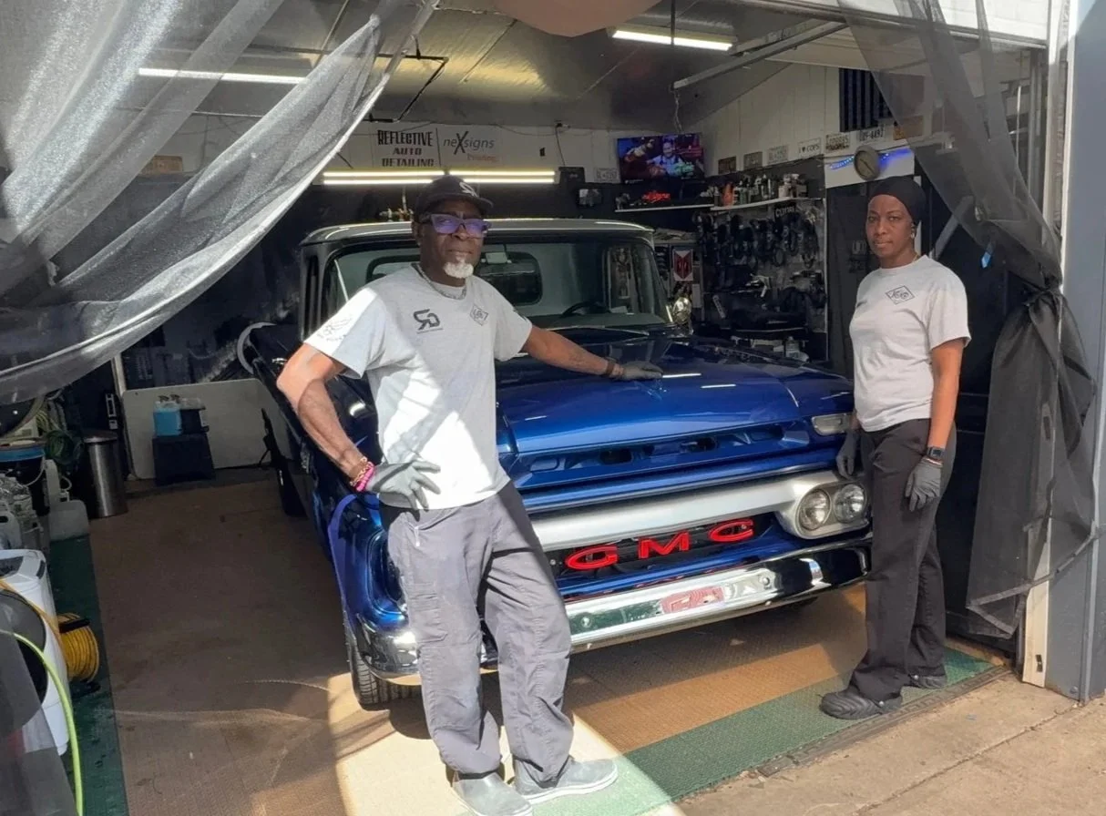 Two people standing in front of a classic blue GMC car inside a garage or workshop.