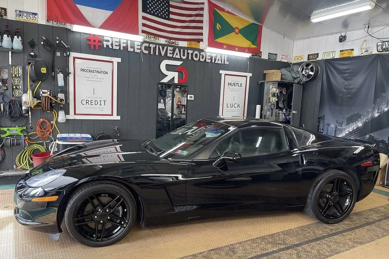 A black sports car parked inside an auto detailing shop with various tools and equipment on the wall, flags hanging from the ceiling, and reflections of lights on the car's surface.