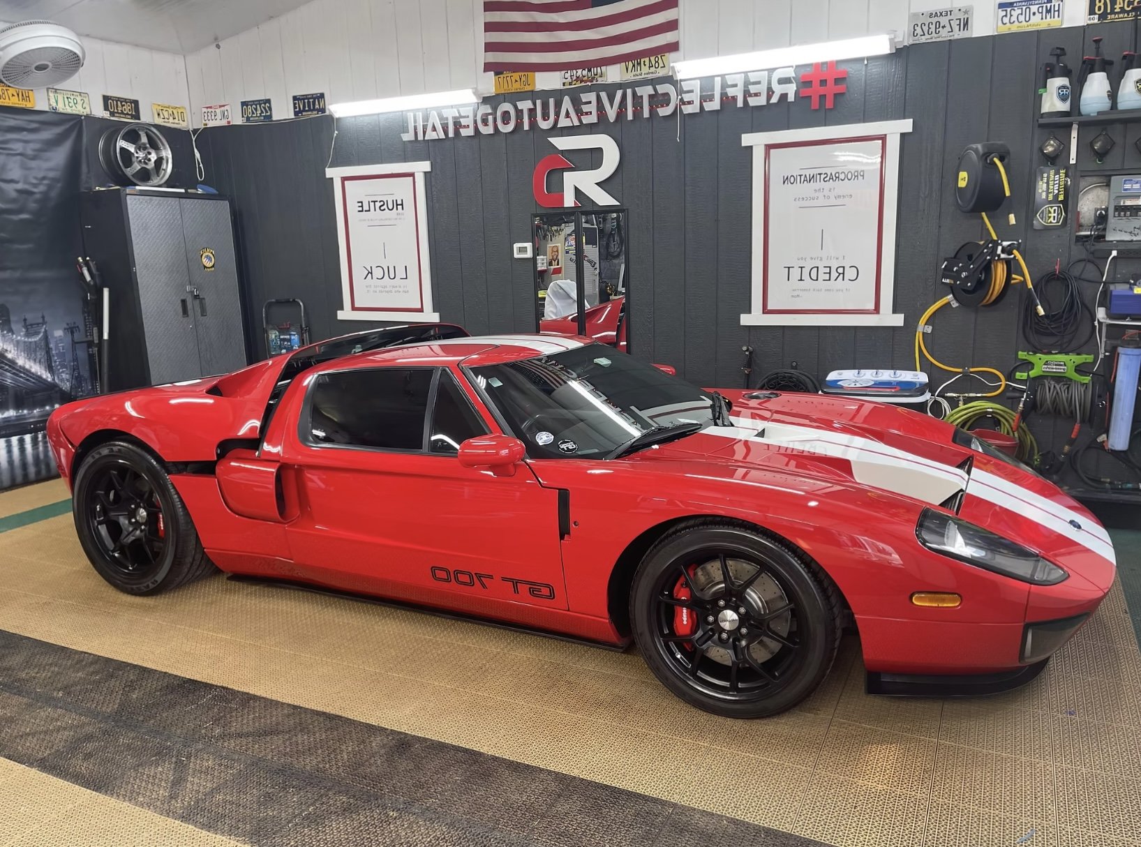 A red Ford GT 700 sports car parked inside a garage with various tools and car accessories on the walls and shelves, including a large gear and a wheel.