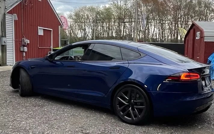 A blue Tesla Model S parked on a gravel lot outside a red building with trees in the background.