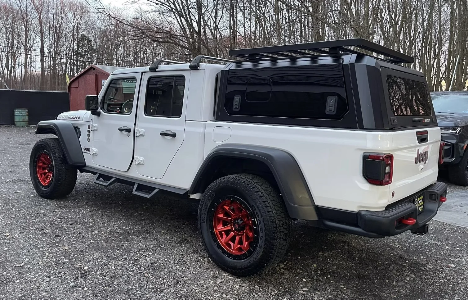 A white Jeep Rubicon pickup truck with red rims parked on a gravel surface near a wooded area and a small red shed.