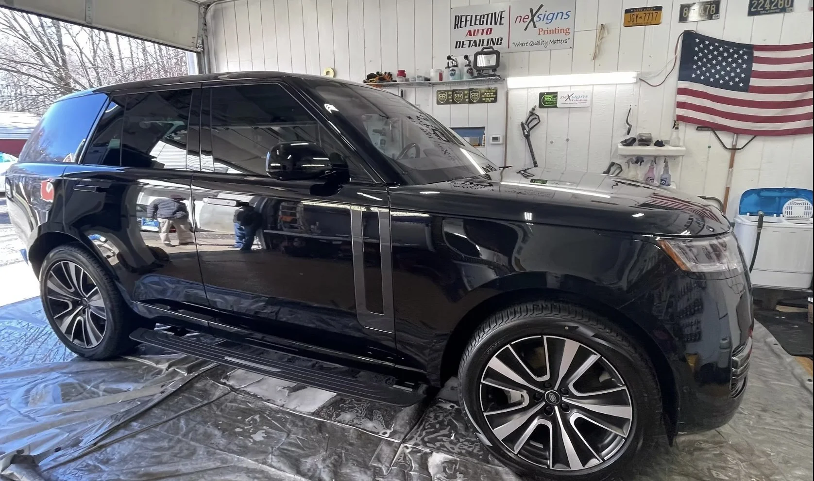 A black Range Rover SUV parked inside a garage with a reflective surface showing the surroundings, including the garage's interior, signs on the wall, American flag, and people taking the photo.