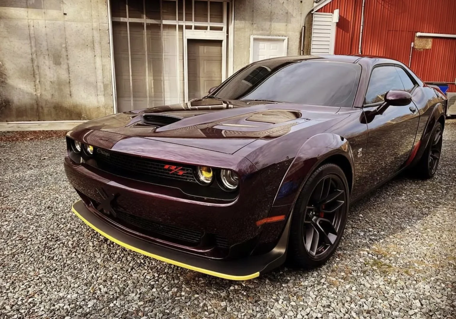 A dark purple Dodge Challenger SRT with yellow accents, parked on gravel outside a building with concrete and red siding. The car has a hood scoop and yellow-tinted headlights.