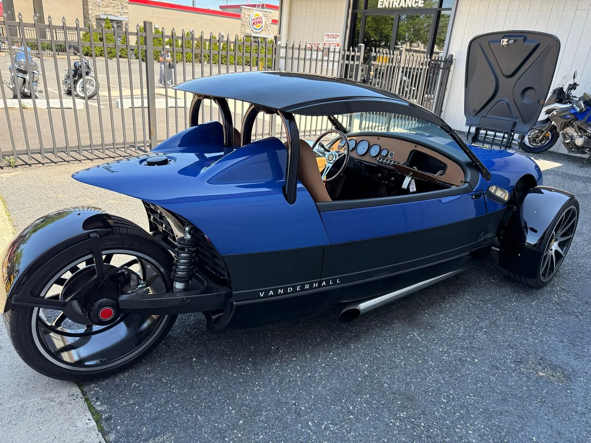 A blue Vanderhall Venice three-wheeled roadster with a tan interior, parked outside a building with a Burger King and other motorcycles in the background.