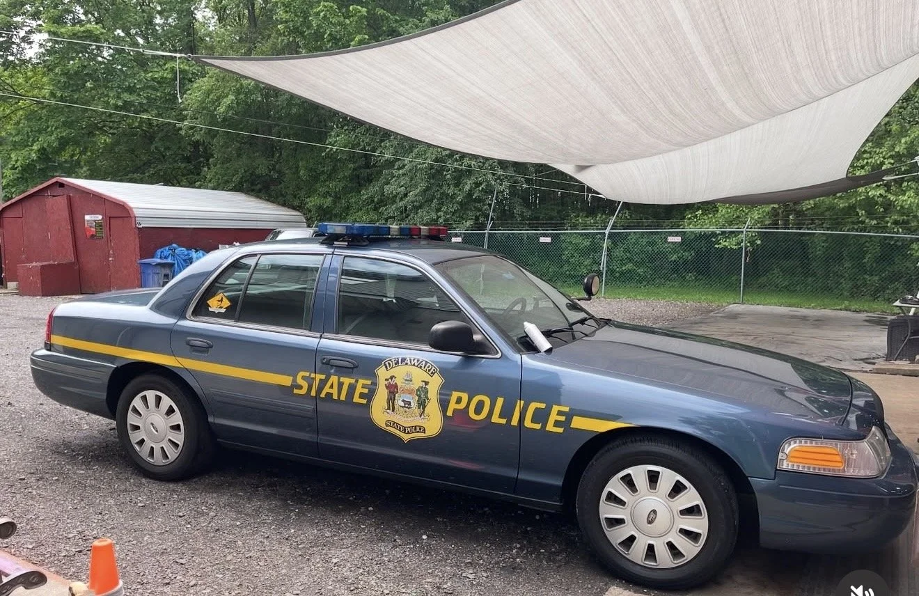 A blue Delaware State Police car parked outdoors on a gravel surface, with a white fabric shade overhead and a red shed in the background.