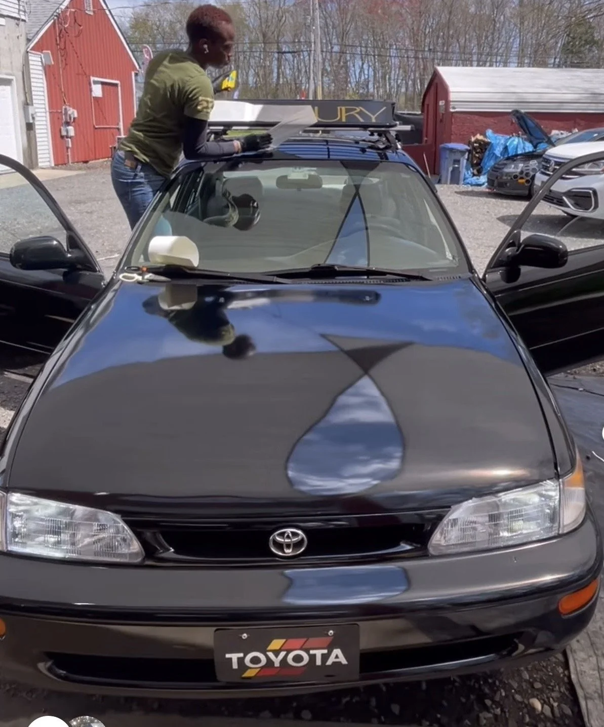 A black Toyota car with open doors parked outdoors, with a woman standing on the roof holding papers, near a red barn and other vehicles in the background.