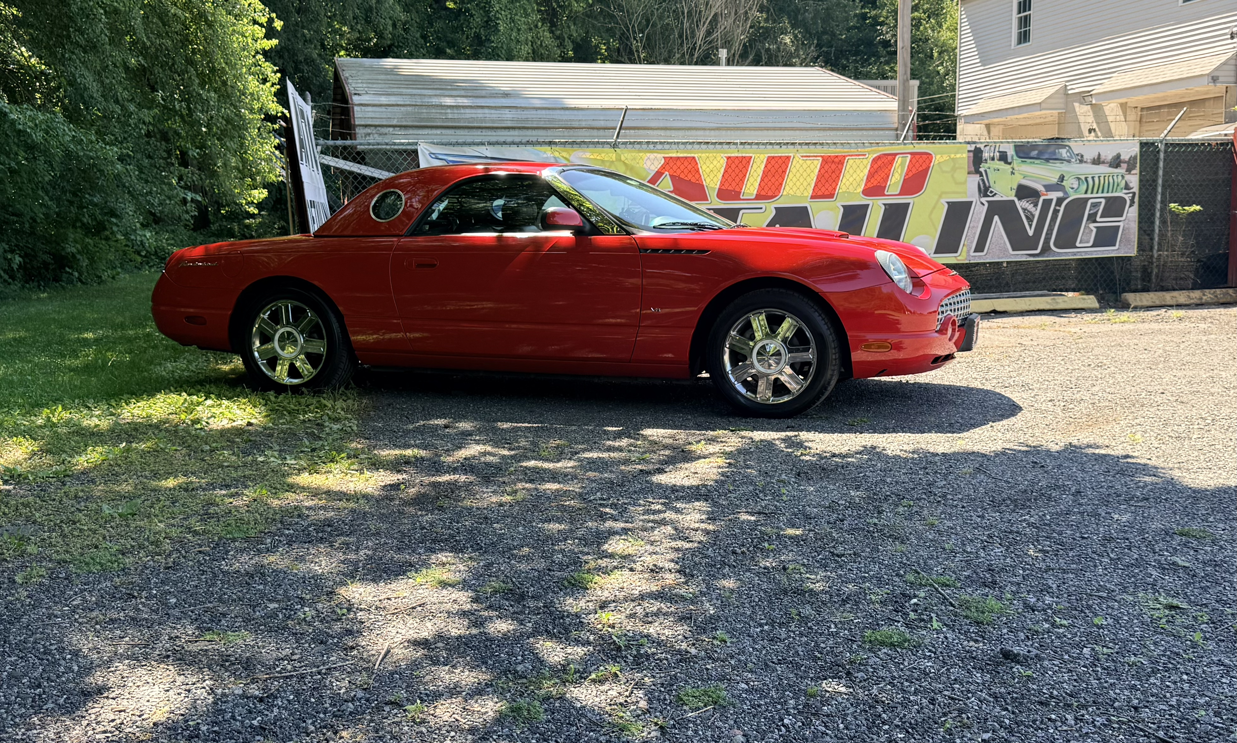 Red sports car parked on gravel driveway in front of a fence with a banner and greenery on the left side and a white house on the right side.