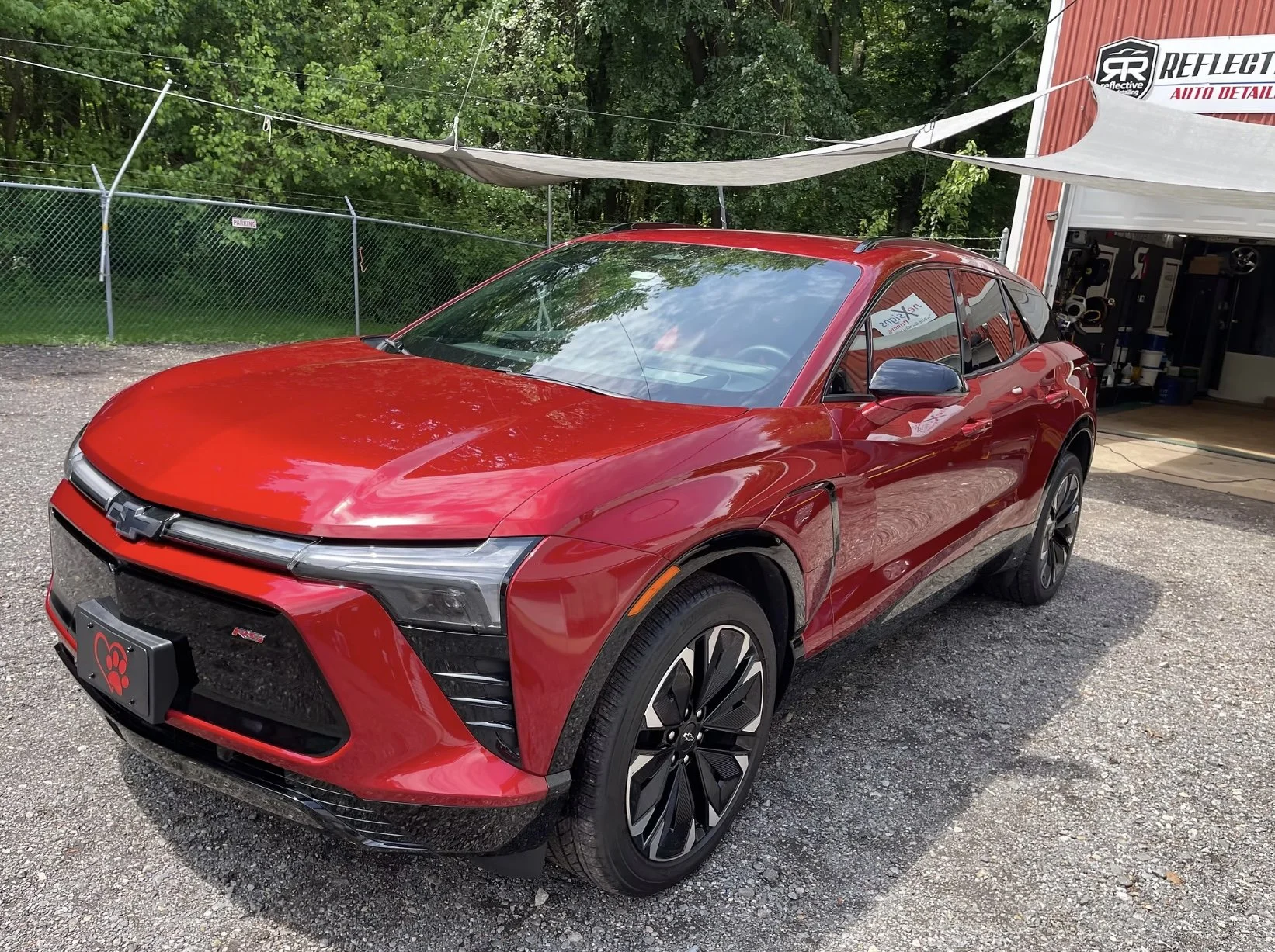Red Chevrolet SUV parked outside a garage with a sign reading Reflective Auto Detailing, surrounded by green trees and a gravel ground.