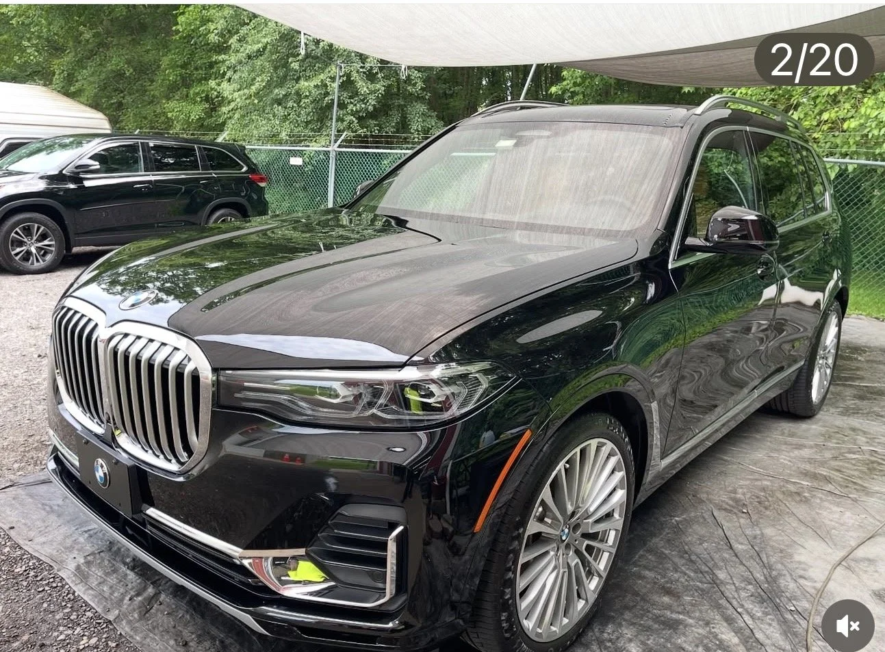 Black BMW SUV parked outdoors under a shade with other vehicles and greenery in the background.