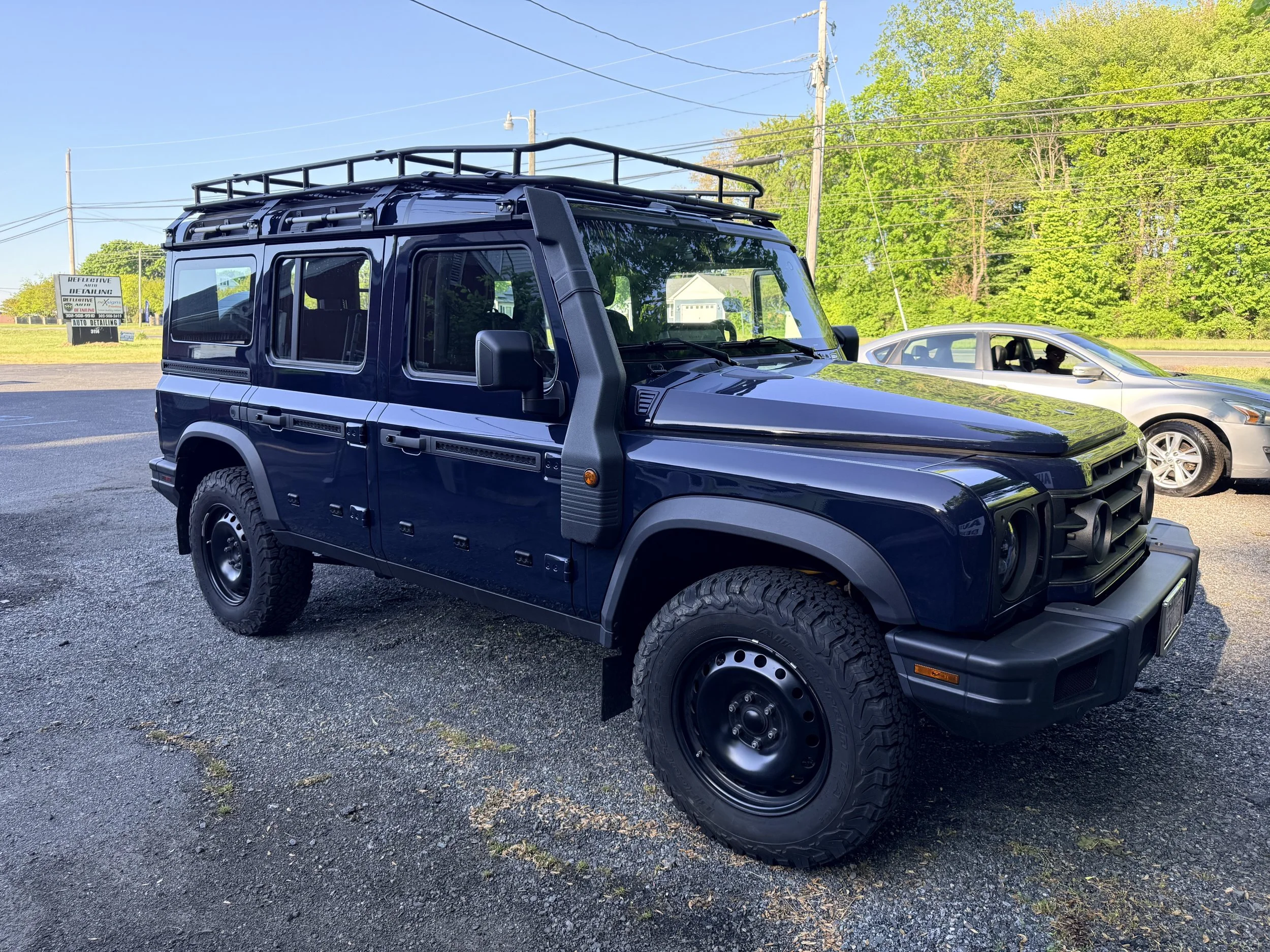 A black off-road vehicle with a roof rack parked on a paved lot, with a silver car and green trees in the background under a clear blue sky.