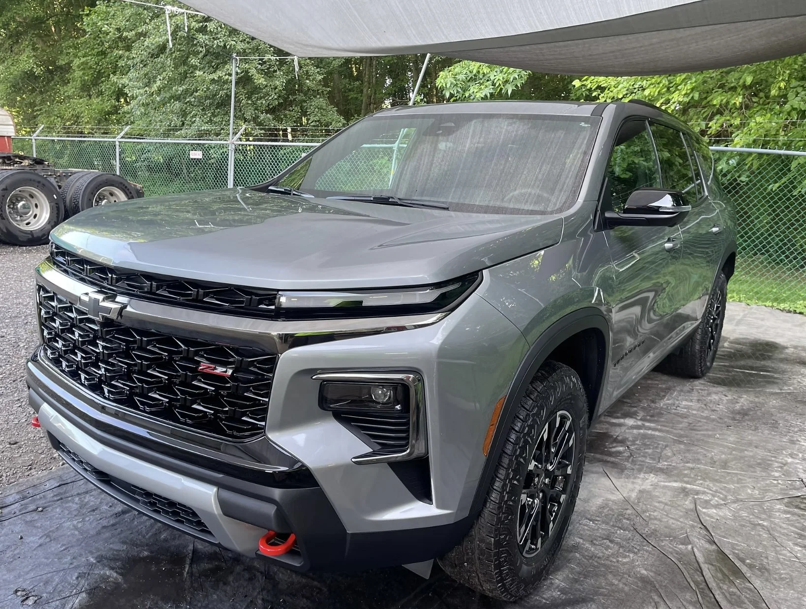 A silver Chevrolet SUV parked under a canopy with a black grille, black wheels, and red tow hooks, surrounded by green trees and a chain-link fence in the background.
