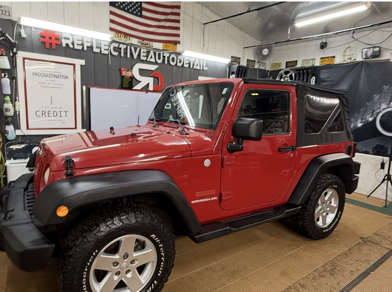 Red Jeep Wrangler with black soft top parked inside a garage or workshop with automotive signs and tools on the walls.