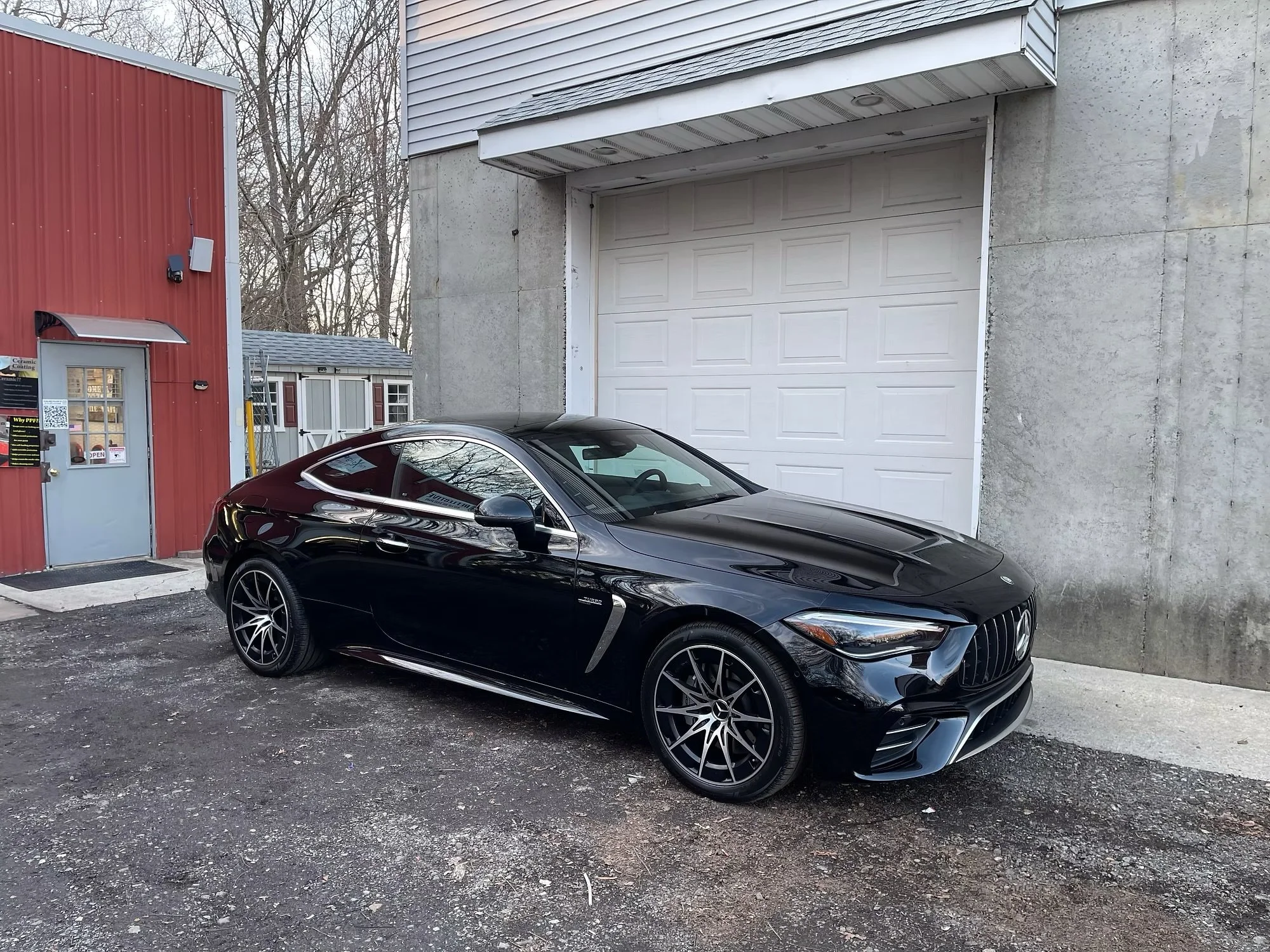 A black Mercedes Benz parked in front of a gray garage door next to a small red building with a door and window. The ground is gravel and there are trees in the background.
