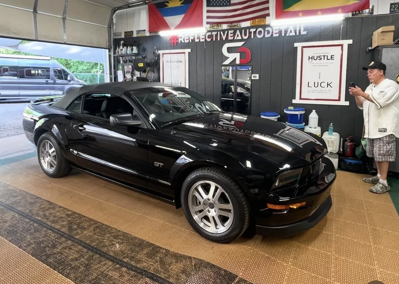 A black Ford Mustang GT convertible parked inside a garage or auto detailing shop. The garage has a black wall with various posters and flags, including an American flag and flags from other countries. There is a person on the right side of the image