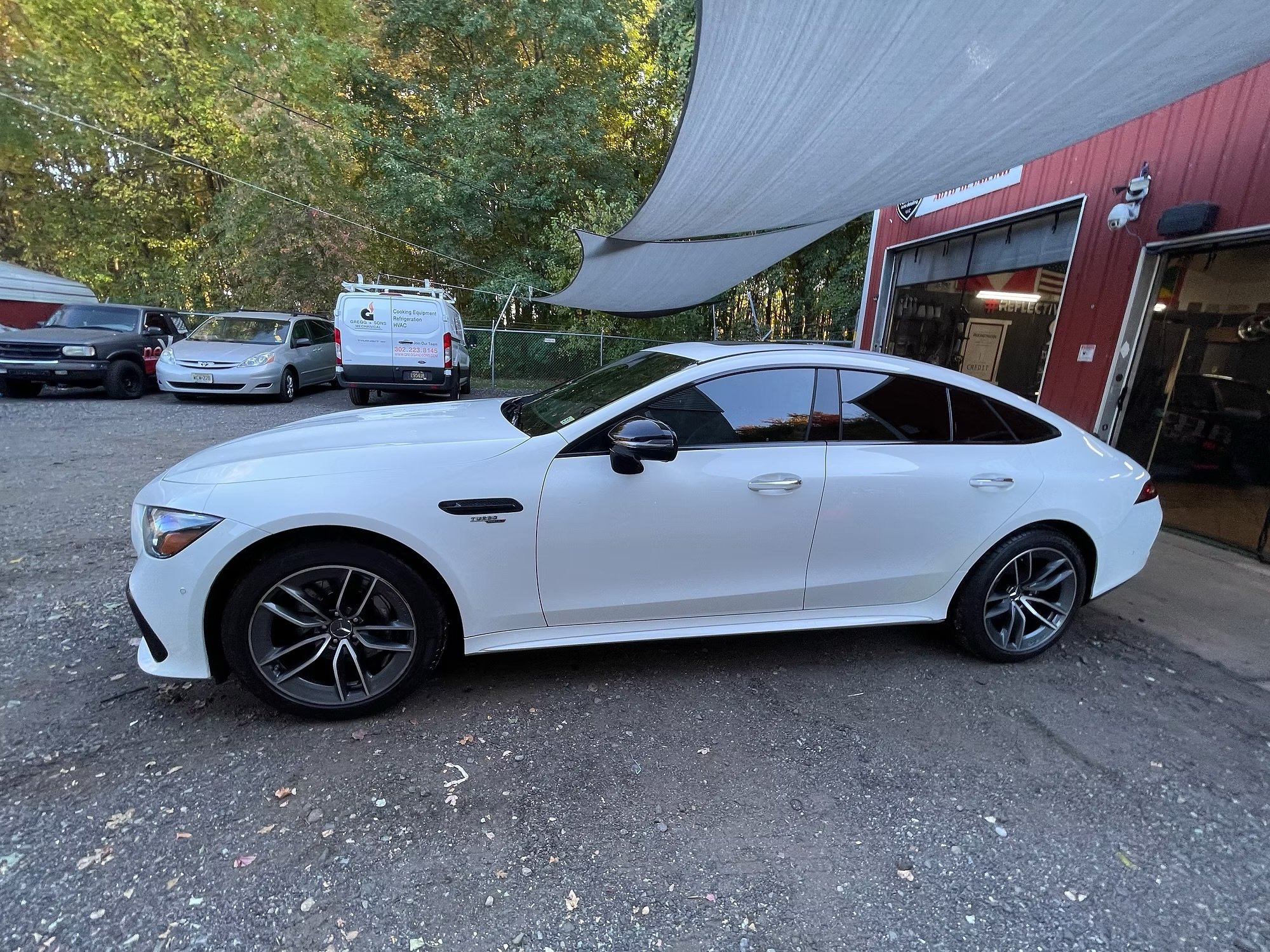 A white sports car parked outdoors on a gravel surface near a red building with an open garage door. Several other vehicles are parked nearby, and a light-colored sunshade extends over the parking lot. There are trees in the background.