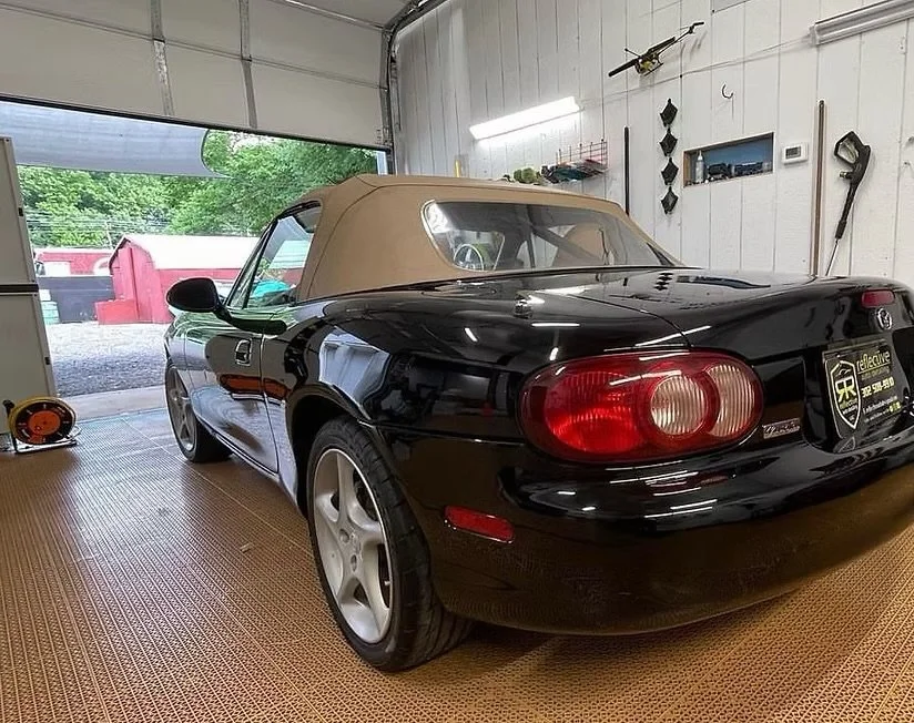 Black Mazda convertible car with a tan soft top parked inside a garage.
