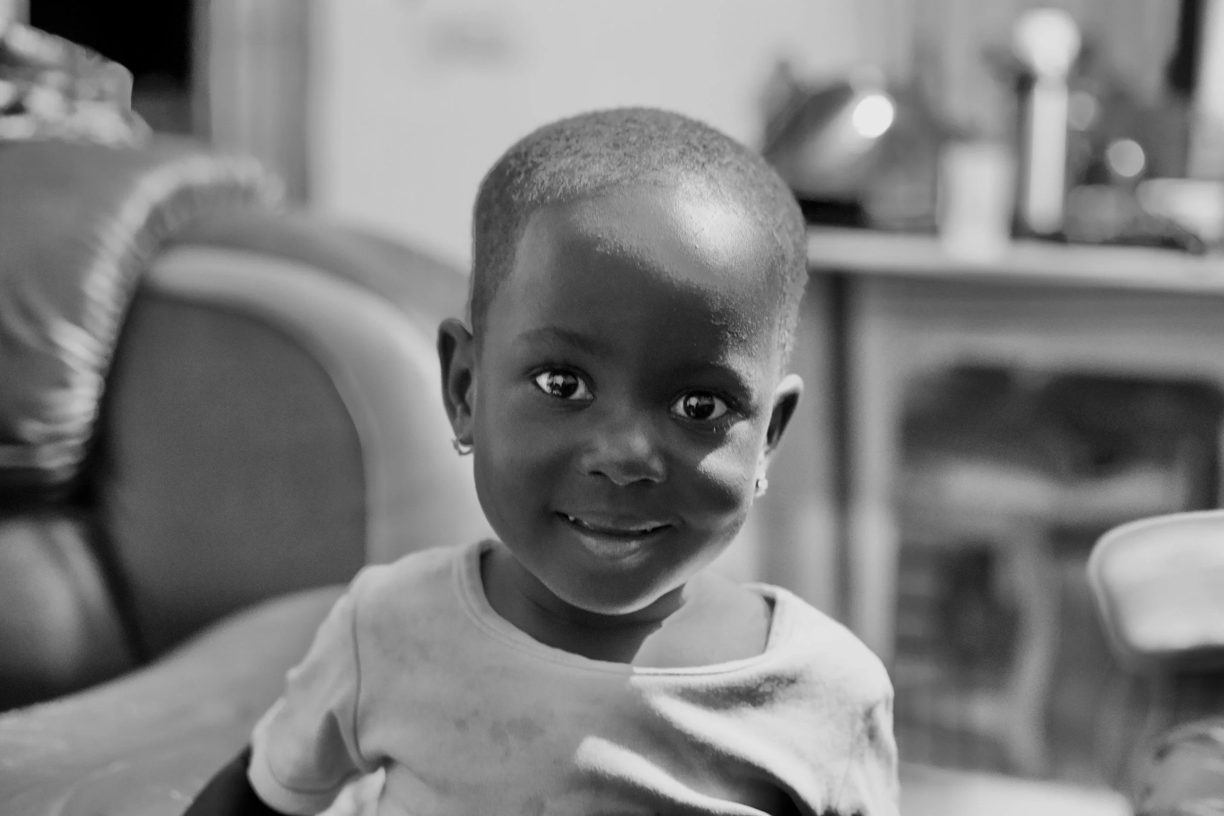 A young African American girl with a short haircut and earrings, smiling at the camera in a home setting in black and white.