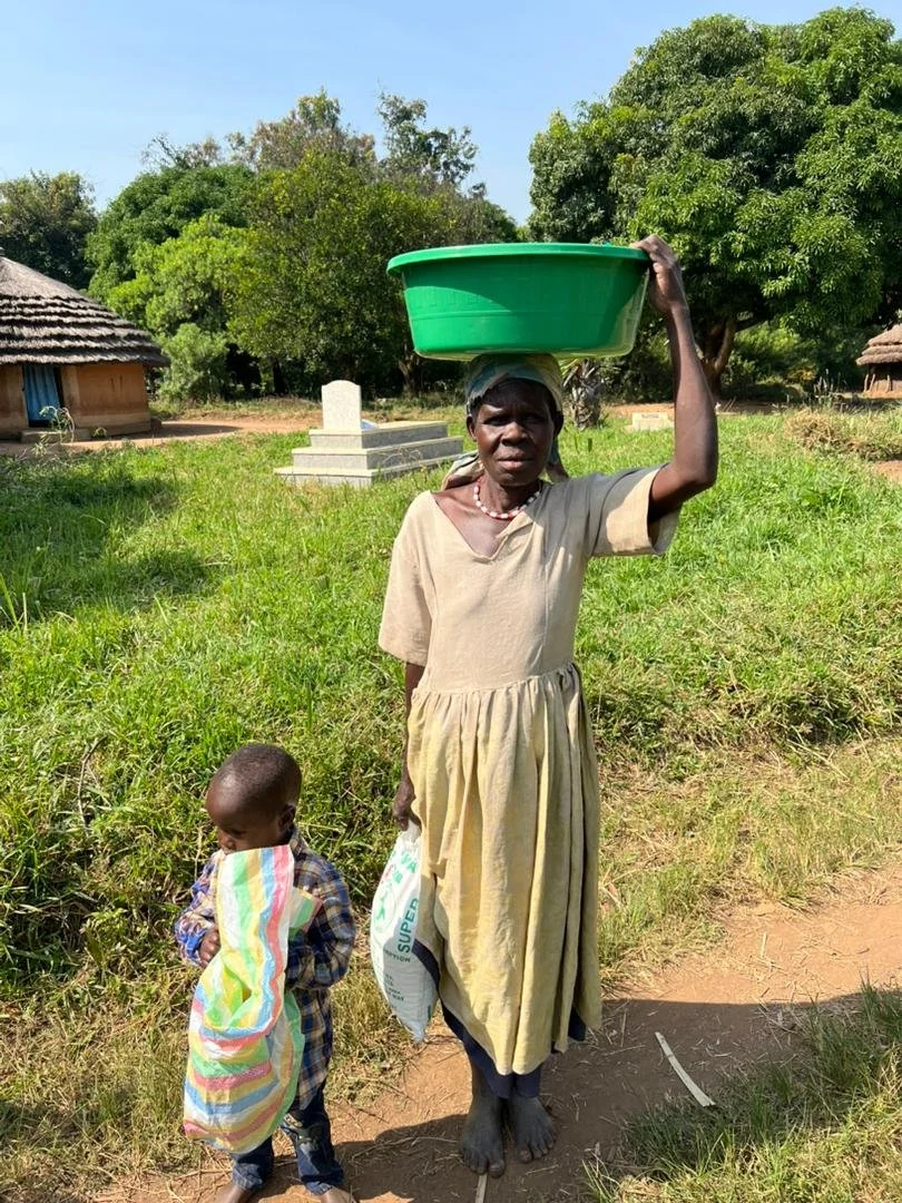 A woman standing outdoors in a rural area, carrying a large green basin on her head, and holding a bag in her left hand. A young child is walking beside her, clutching a colorful striped cloth. There are trees, grass, traditional huts, and a grave in the background.
