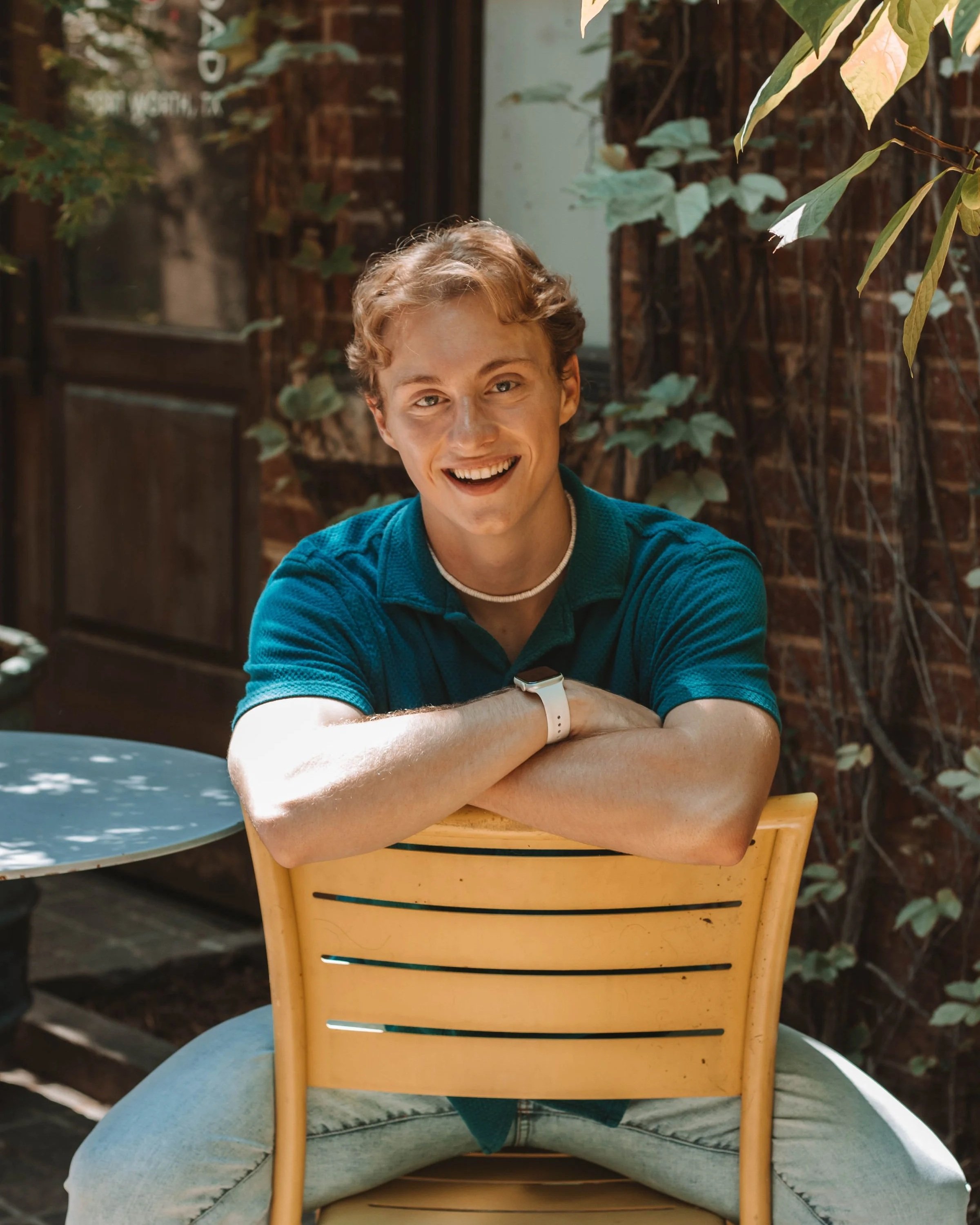 A young man with curly blonde hair smiling, sitting outside behind a yellow chair, wearing a blue shirt, a white choker necklace, and a watch, surrounded by greenery and brick walls.