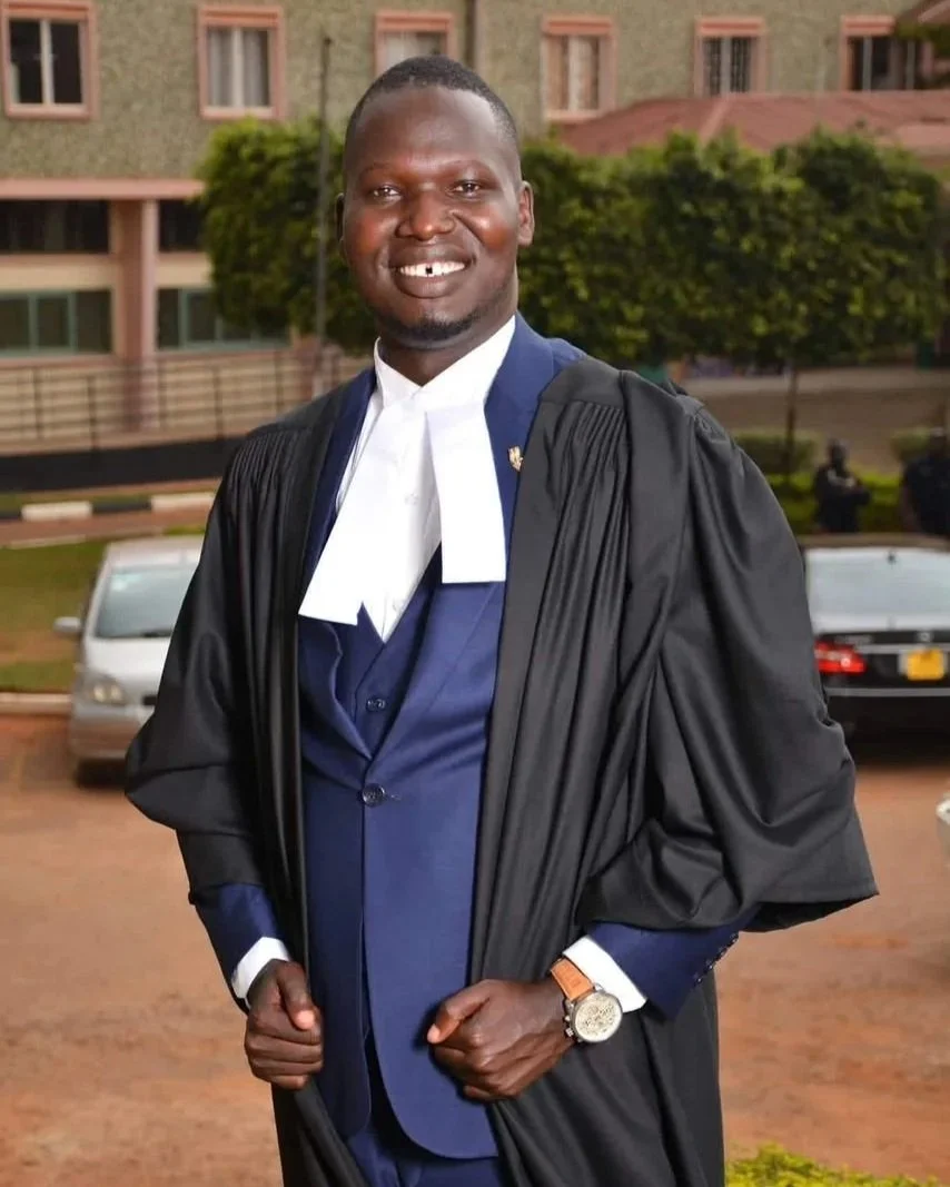 A smiling man wearing academic regalia, including a black gown and a white and blue academic hood, standing outdoors in front of a building with trees and parked cars.
