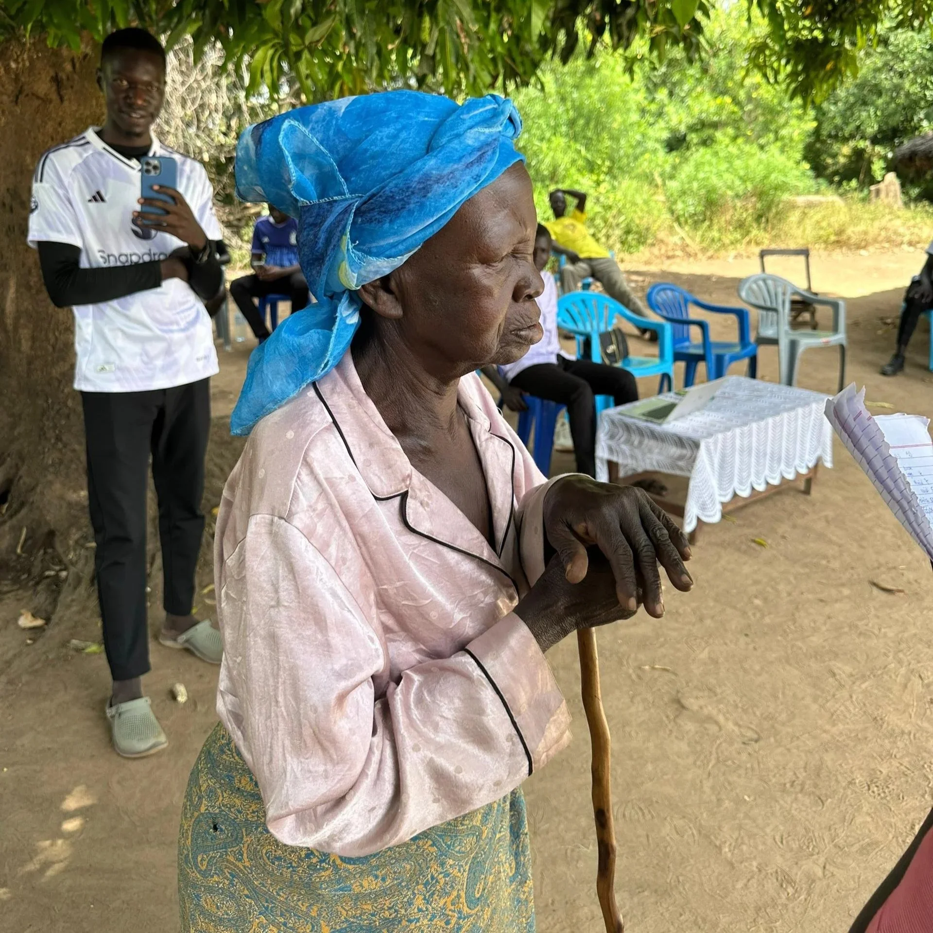 An elderly woman wearing a blue headscarf and pink blouse with black piping, holding a wooden cane, appears to be in a rural outdoor setting with several people sitting and standing around in the background.